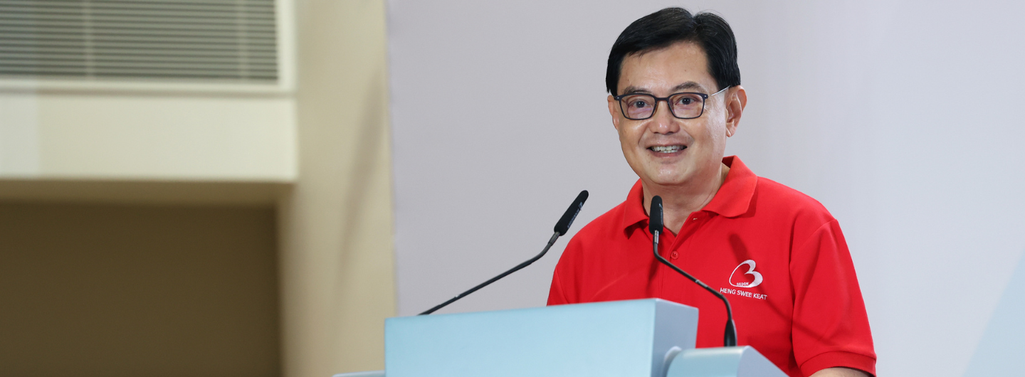 Heng Swee Keat at a light blue podium, wearing glasses and a red polo shirt with a logo.